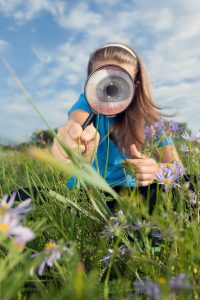 Et barn holder et forstørrelsesglas, hvor igennem man kan se et stort øje, der kikker på græsses og lilla blomster.
